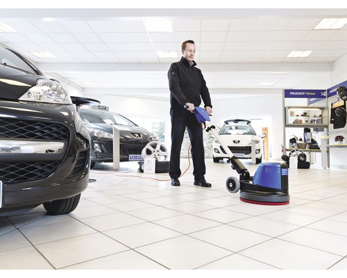Mann reinigt einen Ausstellungsraum mit einer Reinigungsmaschine, Autos sind im Hintergrund zu sehen.