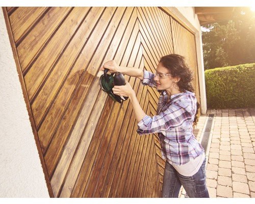 Frau schleift Holztor mit Schleifgerät. Heimwerkerprojekt im Aussenbereich.