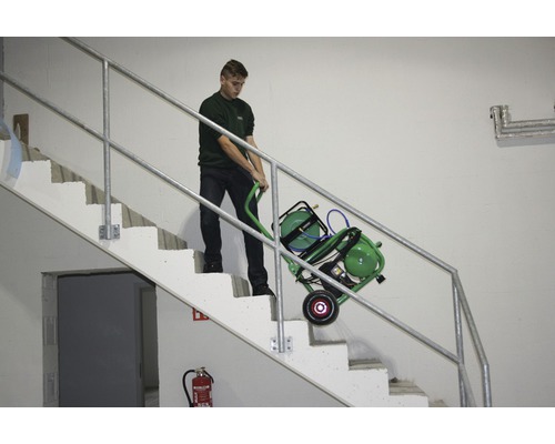 Mann transportiert einen grünen Kompressor mit Schlauchwagen auf einer Treppe.