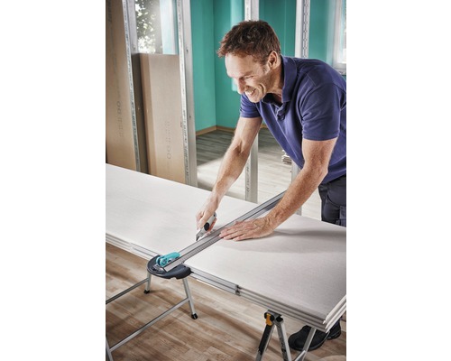 Ein Mann schneidet eine Gipskartonplatte mit einem Cuttermesser und einem Lineal auf einem Tisch.