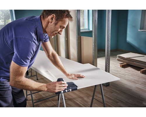 Mann bearbeitet Gipskartonplatte mit Kantenhobel auf einer Werkbank in einem Rohbau.