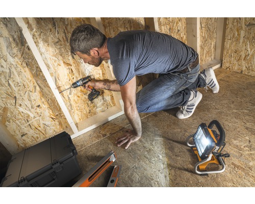 Handwerker arbeitet mit einem Akku-Bohrschrauber in einem Rohbau aus Holz, ausgeleuchtet von einem Baustrahler.