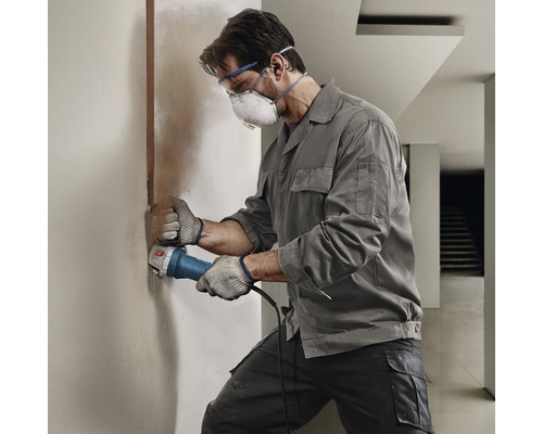 Ein Handwerker fräst mit einer Mauernutfräse eine Nut in eine Wand, er trägt dabei eine Schutzmaske und eine Schutzbrille.