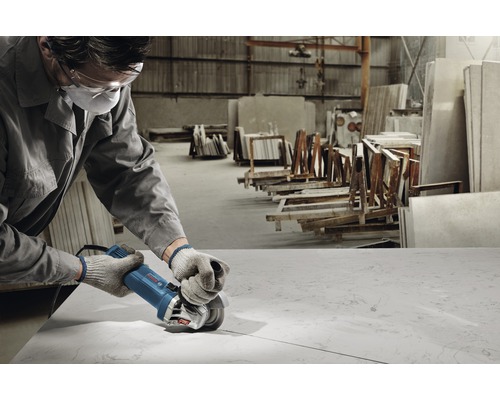 Arbeiter mit Augenschutz und Mundschutz verwendet Winkelschleifer für die Bearbeitung einer Steinplatte in einer Werkstatt.