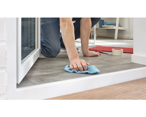 Eine Person kniet und reinigt einen gefliesten Boden mit einem Tuch.