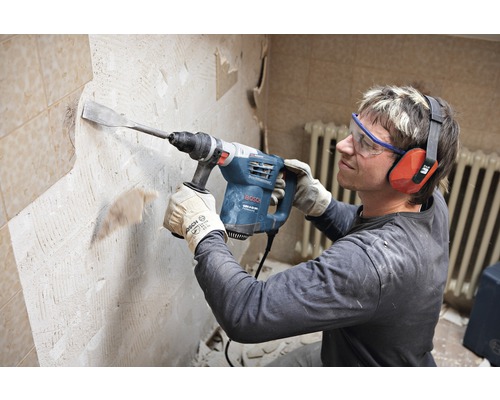 Ein Mann benutzt einen Abbruchhammer zum Entfernen von Fliesen von einer Wand. Er trägt dabei Schutzbrille und Gehörschutz.