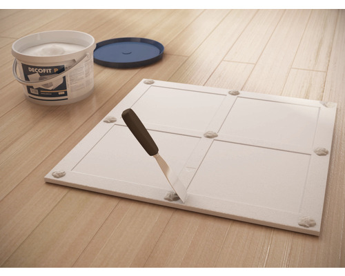 Bild zeigt eine Montageplatte, einen Eimer Klebstoff und einen Spachtel auf einem Holzboden.