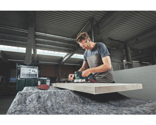 Ein Mann schleift ein Holzbrett mit einem Exzenterschleifer in einer Werkstatt.