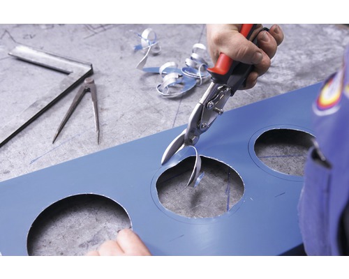 Eine Person schneidet mit einer Blechschere ein Loch in ein blaues Blech. Ein Winkel und ein Zirkel liegen auf dem Tisch.