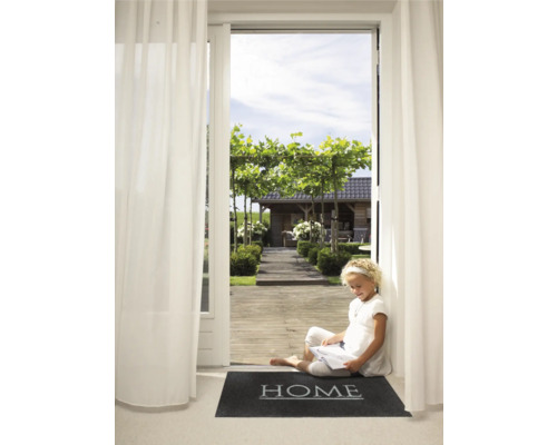 Eine junge Leserin sitzt auf einer Fußmatte mit dem Wort ''Home'' vor einer offenen Terrassentür mit Blick auf einen Garten