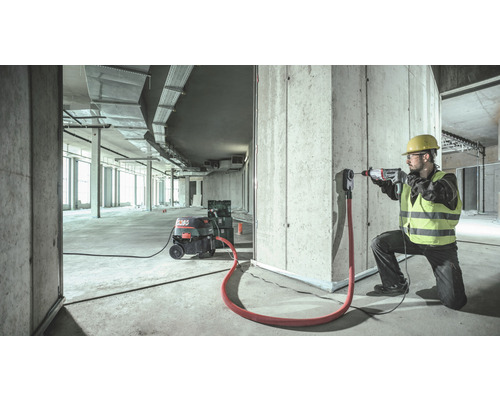 Ein Bauarbeiter mit Schutzhelm und Schutzweste bohrt mit einem Bohrhammer in eine Wand, wobei ein Industriesauger Staub absaugt.