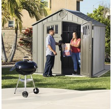 Gartenszene mit Gerätehaus, Grill und zwei Personen, die Holzkohle tragen
