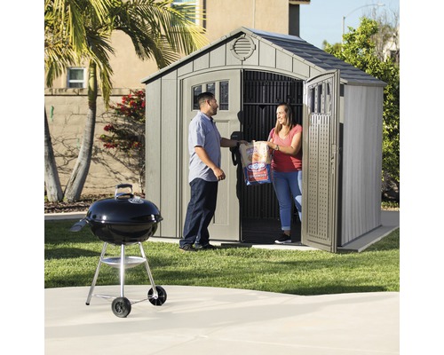 Gartenszene mit Gerätehaus, Grill und zwei Personen, die Holzkohle tragen