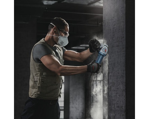 Ein Mann mit Schutzmaske und Schutzbrille arbeitet mit einem Winkelschleifer an einer Betonwand.