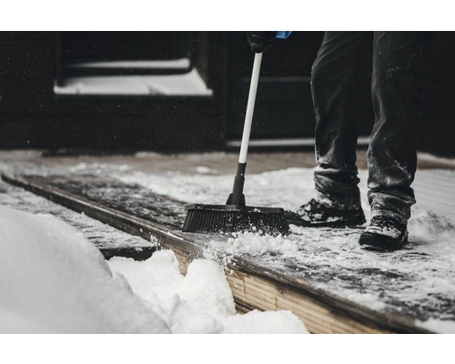 Person kehrt Schnee mit einem Besen von einer Holzterrasse