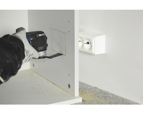 Eine Person verwendet ein Multifunktionswerkzeug, um einen Ausschnitt in ein Holzstück zu schneiden, in der Nähe einer Steckdose.