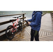 Eine Person reinigt ein Fahrrad mit einem tragbaren Hochdruckreiniger auf einer Holzbrücke in der Nähe eines Gewässers.