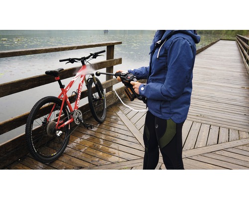 Eine Person reinigt ein Fahrrad mit einem tragbaren Hochdruckreiniger auf einer Holzbrücke in der Nähe eines Gewässers.