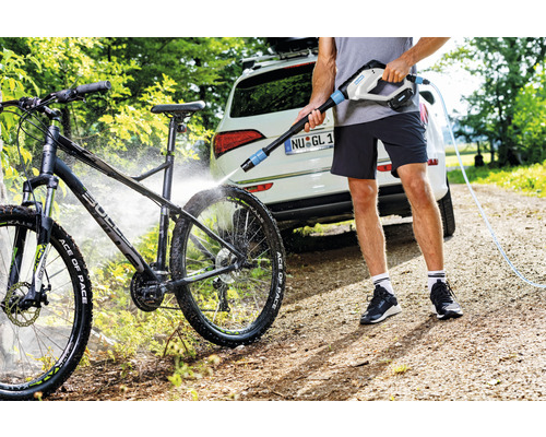 Mann reinigt ein Fahrrad mit einem tragbaren Hochdruckreiniger neben einem Auto.