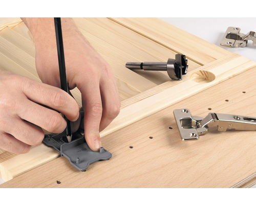 Eine Person markiert die Position für ein Scharnier mit einer Schablone und einem Bleistift auf einem Holzbrett, neben einem Forstnerbohrer und einem Scharnier.