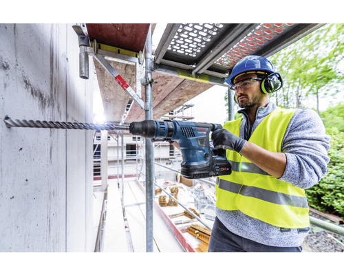 Ein Mann bohrt mit einem Bohrhammer ein Loch in eine Betonwand. Er trägt Schutzkleidung.
