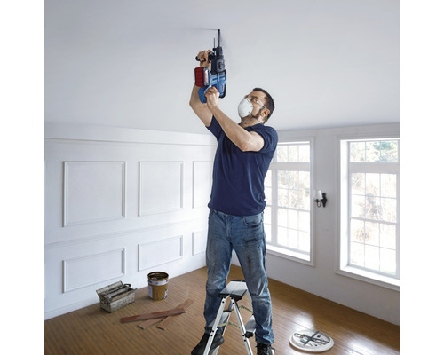 Mann bohrt mit einer Bohrmaschine ein Loch in die Decke stehend auf einer Leiter in einem Raum.