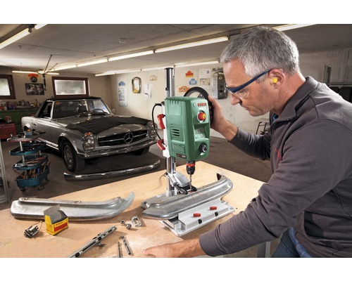 Werkstattszenario mit einer Bosch Tischbohrmaschine beim Bohren eines Werkstücks, ein Mann trägt dabei Schutzbrille und Gehörschutz.