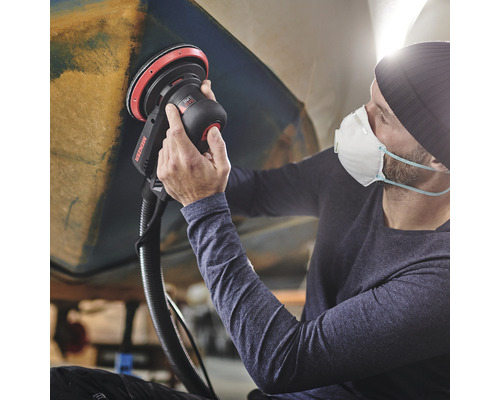 Ein Mann schleift ein Objekt mit einem Exzenterschleifer der Marke Menzer. Er trägt eine Atemschutzmaske.