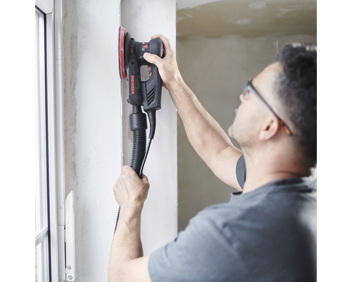 Mann schleift mit einem Exzenterschleifer eine Wand. Das Gerät ist mit einem Absaugschlauch ausgestattet.
