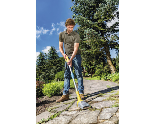 Ein Mann benutzt einen Fugenreiniger auf einem gepflasterten Weg im Garten, um Unkraut zu entfernen.
