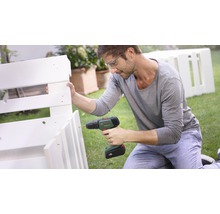 Mann mit Schutzbrille, der eine Bohrmaschine benutzt, um ein weißes Holzregal im Garten zu montieren. Bosch Logo.