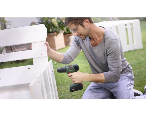 Mann mit Schutzbrille, der eine Bohrmaschine benutzt, um ein weißes Holzregal im Garten zu montieren. Bosch Logo.