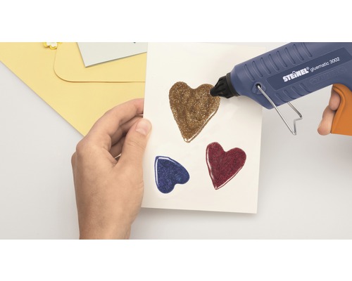 Heißklebepistole beim Aufbringen von Glitzerherzen auf Papier