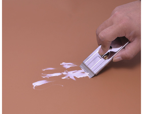 Eine Hand benutzt einen Glasschaber, um Farbe von einer Oberfläche zu entfernen.