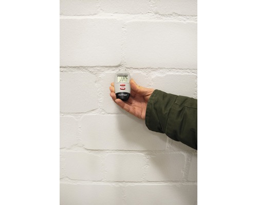 Eine Hand hält ein Feuchtigkeitsmessgerät vor einer Wand.