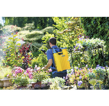 Ein Mann verwendet eine Rückenspritze zur Gartenarbeit inmitten von Blumen und Pflanzen.