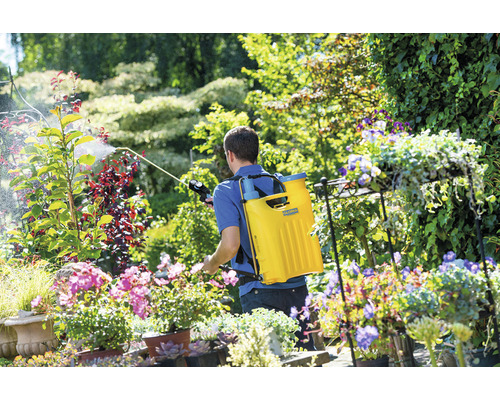 Ein Mann verwendet eine Rückenspritze zur Gartenarbeit inmitten von Blumen und Pflanzen.