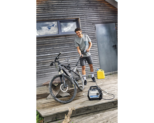 Mann reinigt Fahrrad mit einem Hochdruckreiniger auf einer Holzterrasse vor einem Haus.