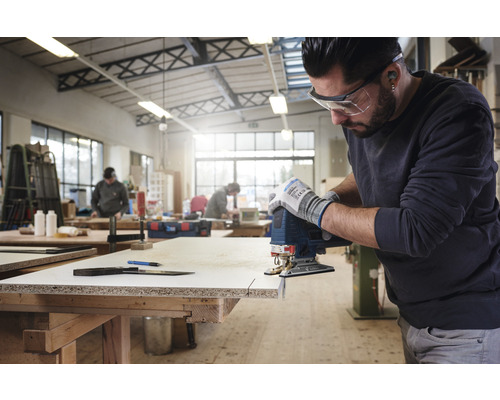 Ein Mann benutzt eine Stichsäge, um ein Holzbrett in einer Werkstatt zu schneiden, wobei er Schutzbrille und Handschuhe trägt.