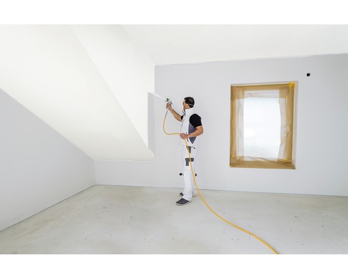 Ein Mann streicht einen Raum mit einer Farbspritzpistole, Fenster sind abgedeckt.