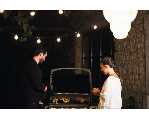Ein Mann und eine Frau grillen Fleisch und Kartoffeln auf einem Grill im Freien, beleuchtet mit einer Lichterkette.