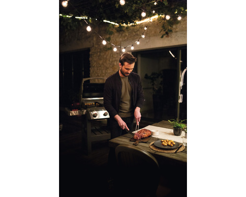 Mann schneidet Fleisch auf einer Terrasse mit Grill und Lichterkette