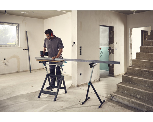 Ein Mann schneidet ein Metallprofil auf einer Werkbank in einem unrenovierten Raum.