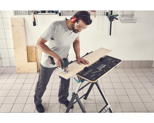 Ein Mann sägt mit einer Stichsäge ein Holzbrett auf einem Werktisch. Er trägt eine Schutzbrille und einen Gehörschutz.