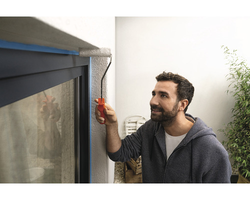 Ein Mann streicht mit einer Farbwalze einen Fensterrahmen, der mit Klebeband abgeklebt ist.