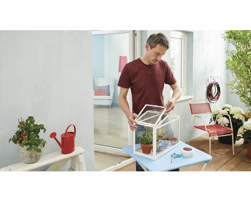 Mann öffnet ein Mini-Gewächshaus auf einem Balkon mit Pflanzen