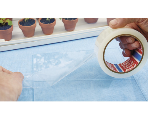 Eine Rolle Tesa Film wird verwendet, um eine Folie zu befestigen. Im Hintergrund sind kleine Pflanztöpfe zu sehen.