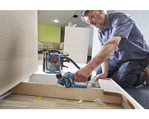 Mann hobelt Holz mit einem Elektrohobel in einer Werkstatt. Ein Staubsauger steht im Hintergrund.