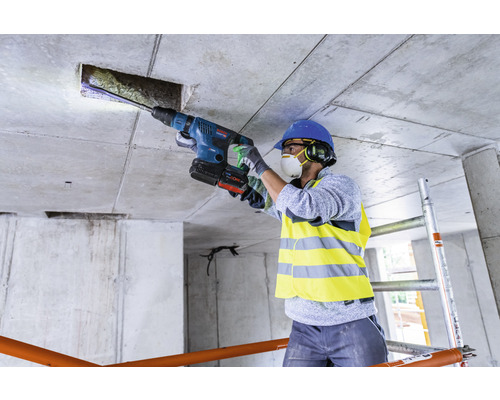 Ein Bauarbeiter bohrt mit einem Bohrhammer ein Loch in eine Betondecke. Der Arbeiter trägt Schutzkleidung.