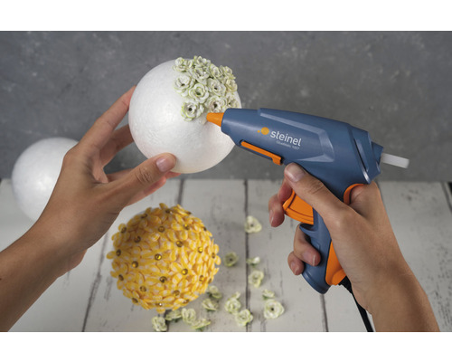 Eine Person dekoriert einen Styroporball mit einer Heissklebepistole und kleinen Blumen.